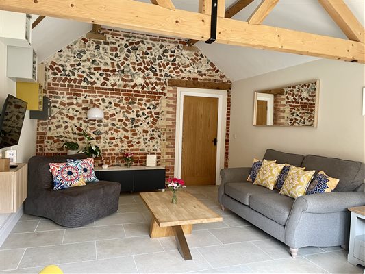 Grey sofa on your right with yellow cushions. Dark grey chair on the left with bright coloured cushions.  Original flint wall at the back with a wooden door to the right.